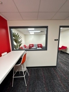 Photo inside of lounge, looking through clear window into study room. White countertop with red stool on lefthand side.