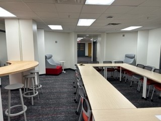 Photo looking from West door into lounge. Light wood countertop and swivel stools on left side, large rectangular table with matching grey chairs on right side. Red and grey armchair in background next to hallway.