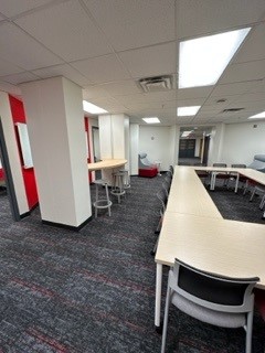 Photo of Student Lounge taken from West entrance. Long wood table with grey chairs, red accent wall and wooden countertop on left-hand side.