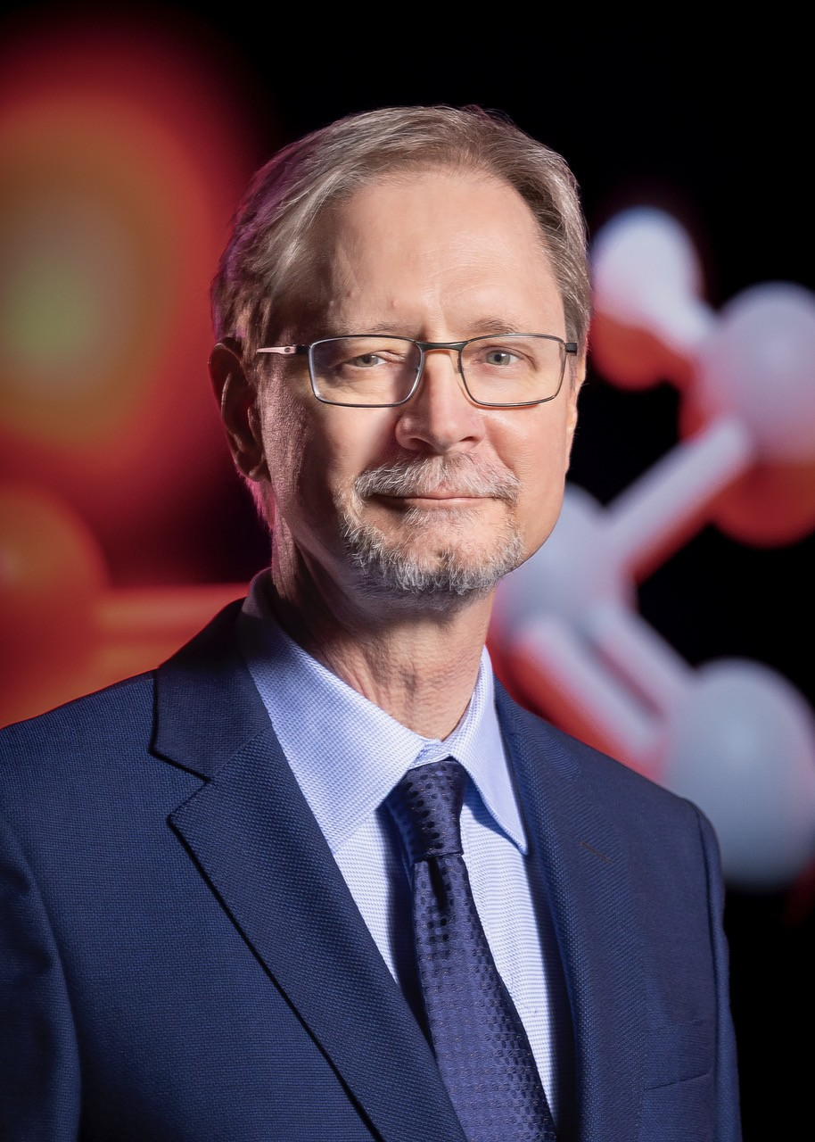 Kenneth Schafer in a blue suit with large graphic of an atom behind him