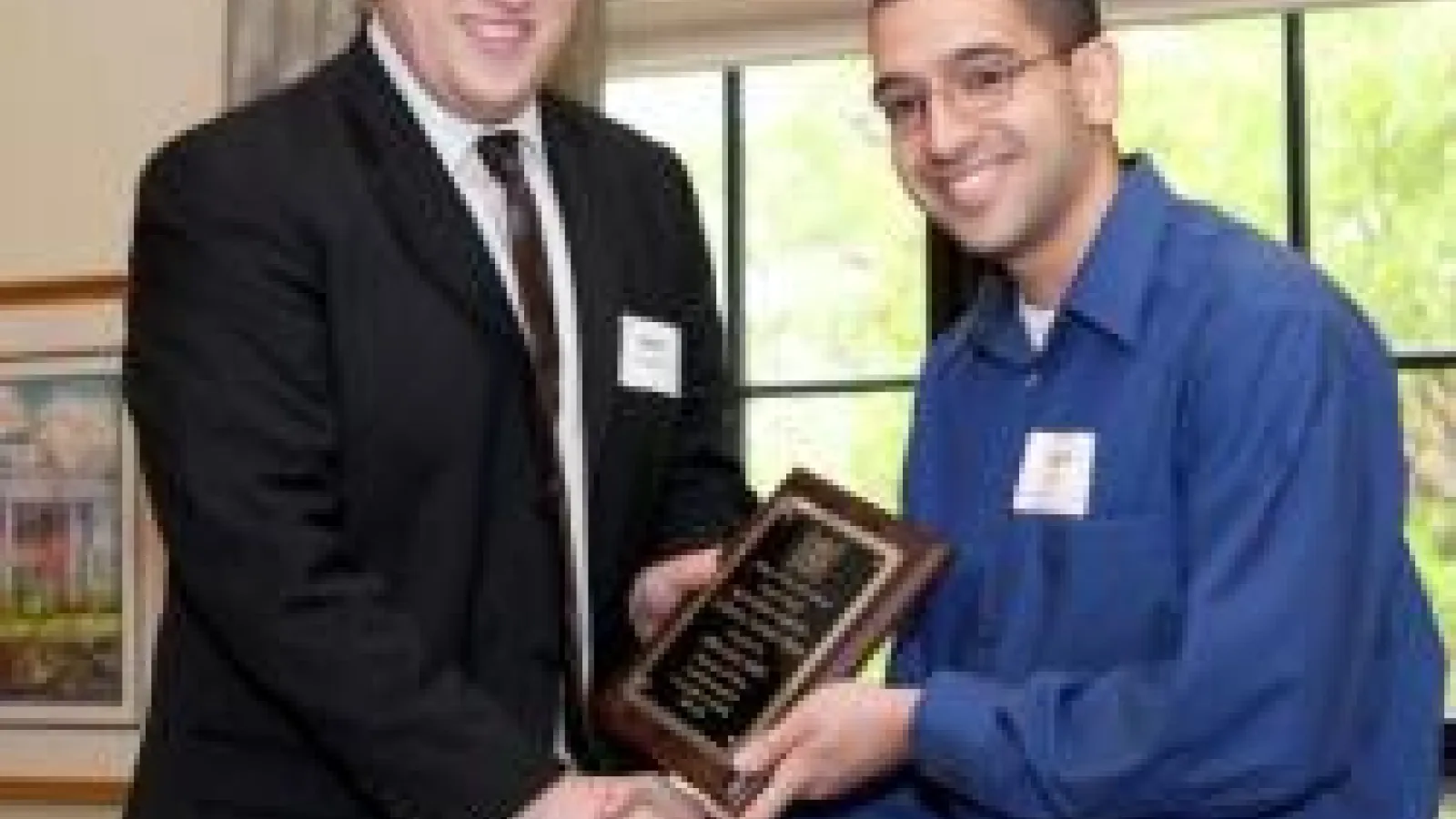 Adam Ahmed shaking hands with the person giving his award to him. They are both holding the plaque in the center.