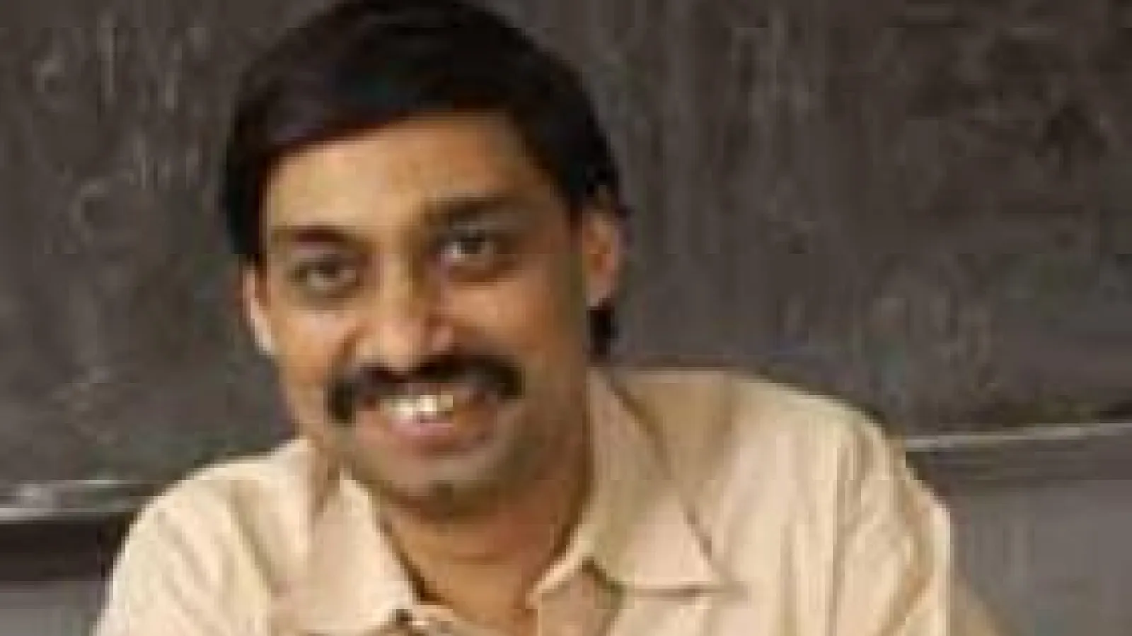 Samir Mathur smiling and sitting at his desk in front of a chalkboard