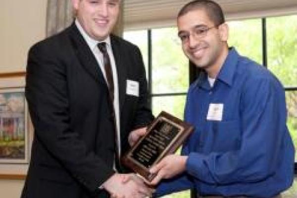 A picture of Adam Ahmed shaking hands with the person giving his award to him. They are both holding the plaque in the center.