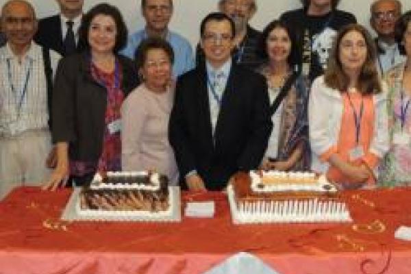 The photo shows the Council at the 50th anniversary cake-cutting ceremony. There are 17 people posing behind a table that had two different cakes on it.