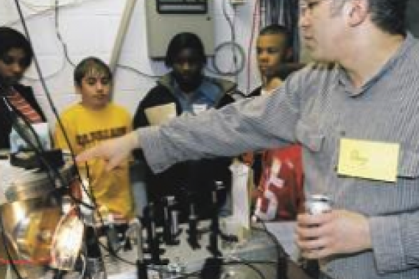 The picture shows Douglass Schumacher working with a group of students in a lab. He is explaining the equipment as the students listen attentively.