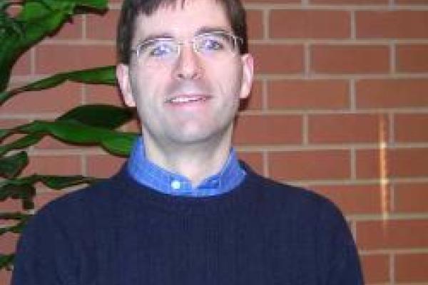 A picture of Bradley Ambrose with a brick wall and tall green plant in the background.