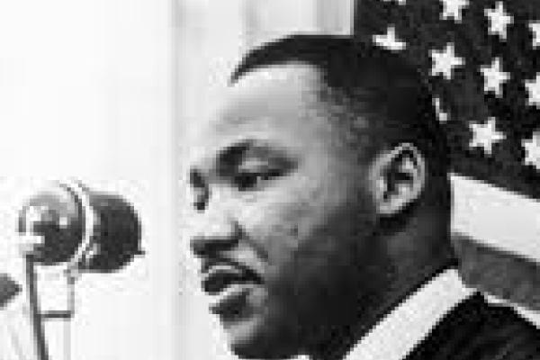 Black and white photo of Martin Luther King Jr Speaking into microphone with American flag in the background
