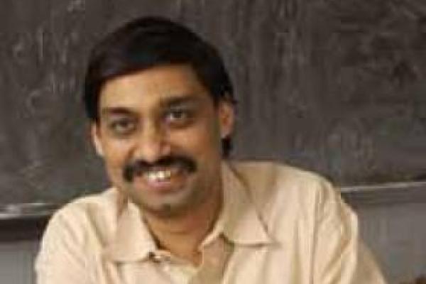 Samir Mathur smiling and sitting at his desk in front of a chalkboard